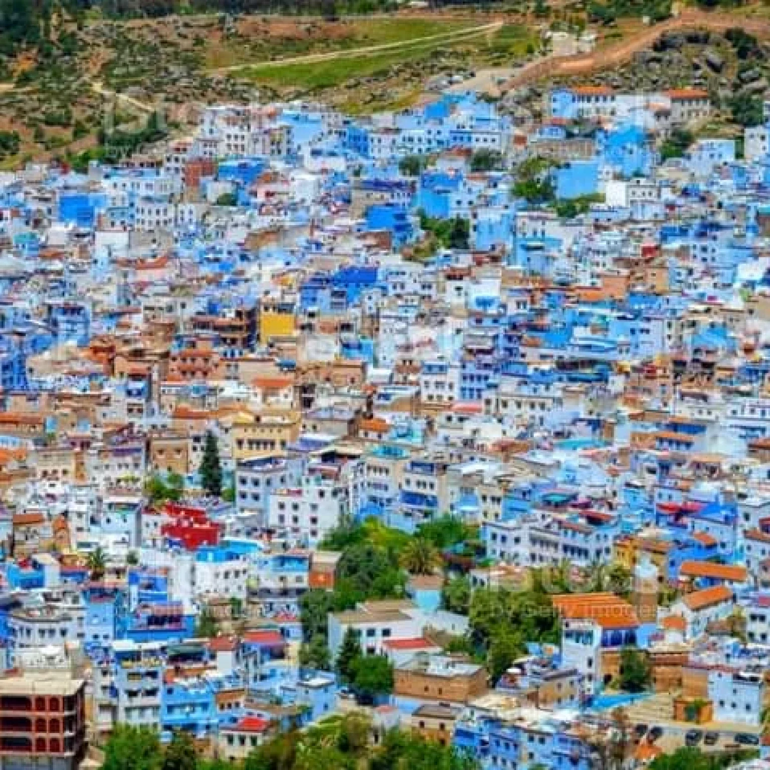 Aerial view of Chefchaouen, Morocco, nestled among rolling hills, showcasing the city's breathtaking beauty with its iconic blue buildings and surrounding verdant landscape.