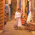 Woman in white blouse and skirt walking amidst vibrant, colorful traditional rugs and textiles displayed on walls and piles in a Marrakech souk, showcasing the rich culture and craftsmanship of Moroccan markets.